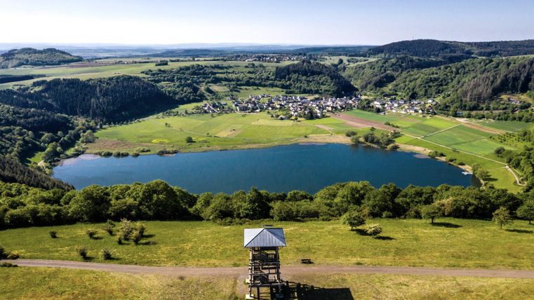 Eine malerische Landschaft mit einem klaren See und üppigen Wäldern. Im Vordergrund steht ein Aussichtsturm, der einen wunderschönen Blick auf die Umgebung bietet.