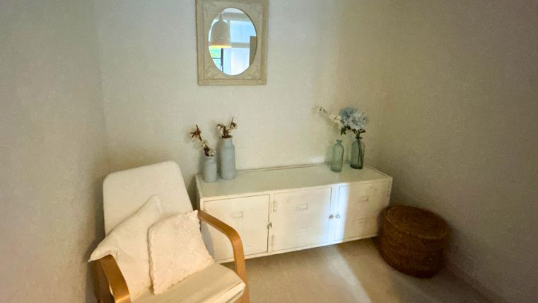 Ein heller Raum mit einem gemütlichen Stuhl und einem kleinen Tisch. Auf dem Tisch stehen Vasen mit Blumen und ein Spiegel hängen an der Wand.