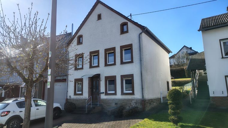 Ein freistehendes Haus mit weißen Wänden und braunen Fensterrahmen. Vor dem Haus befindet sich eine grüne Wiese und ein Gehweg.