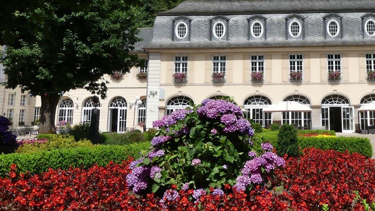 Ein schönes Blumenbeet mit roten und lila Blumen vor einem eleganten Gebäude. Die Umgebung ist grün und einladend.
