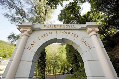 Ein schöner Torbogen mit der Aufschrift "Römerkessel" und "Das Leben erwartet Dich". Umgeben von grünen Bäumen und einer malerischen Landschaft.