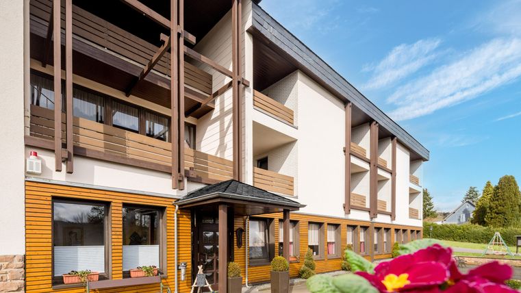 Ein modernes Gebäude mit Holzverkleidung und mehreren Balkonen. Im Vordergrund sind Blumen und eine einladende Eingangstür zu sehen.