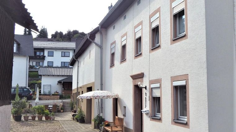 Ein ruhiger Innenhof mit zwei Gebäuden. Auf der rechten Seite steht ein Tisch mit einem Sonnenschirm und einige Pflanzen sind sichtbar.