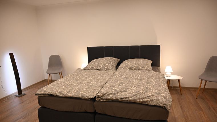 Ein gemütliches Schlafzimmer mit einem Doppelbett und modernen Stühlen. Die Wände sind neutral, und es gibt eine angenehme Beleuchtung.