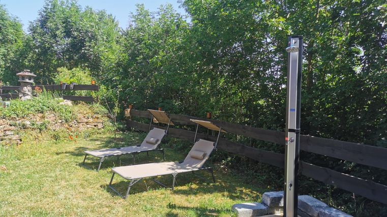 Ein ruhiger Gartenbereich mit zwei Liegen und einer Außendusche. Umgeben von Bäumen und einer Holzlattenzaun.