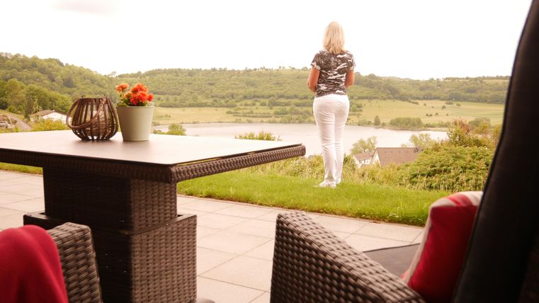 Eine Frau steht auf einer Terrasse und schaut auf eine grüne Landschaft mit einem Gewässer im Hintergrund. Der Tisch ist mit einer Blumenvase und einem dekorativen Korb geschmückt.