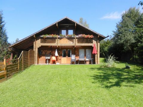 Ein gemütliches Holzhaus mit einem großen Garten. Der Balkon ist mit Blumen und einem Sonnenschirm dekoriert.