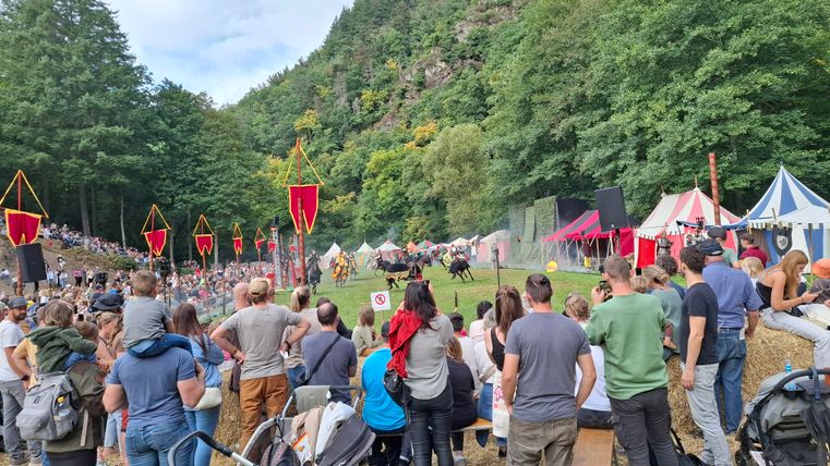Eine große Menschenmenge ist auf einer Wiese versammelt, um eine Veranstaltung im Freien zu beobachten. Im Hintergrund sind historische Zelte und eine bewaldete Landschaft zu sehen.