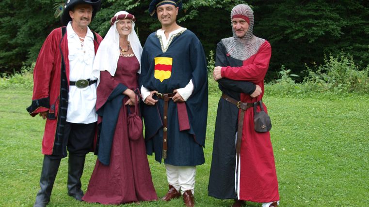 Eine Gruppe von vier Personen in historischen Kostümen steht auf einer grünen Wiese. Sie tragen Kleidung aus verschiedenen Epochen, darunter Röcke, Tuniken und Hüte.