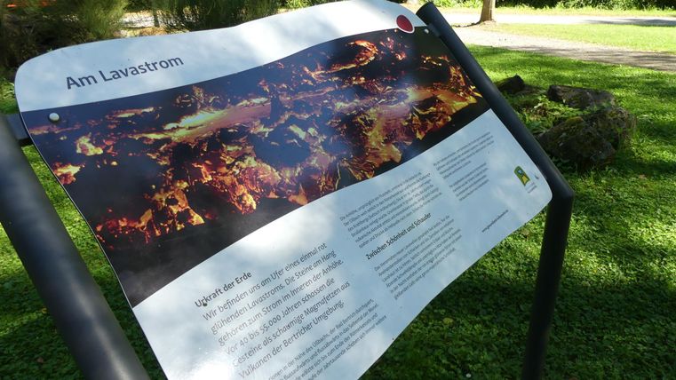 Ein Informationsschild über einen Lavastrom in einem Park. Die Umgebung ist grün mit Bäumen und einer angenehmen Atmosphäre.