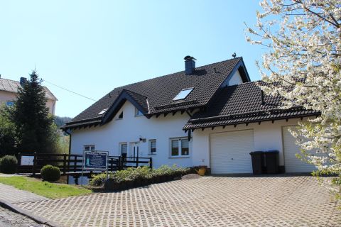 Ein modernes Haus mit einem dunklen Dach und einer Auffahrt. Bäume und Sträucher umgeben das Gebäude in einer ruhigen Umgebung.