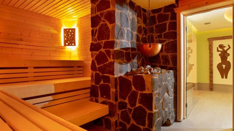 Eine moderne Sauna mit Holzsitzbänken und einem Steinofen. Der Raum ist warm beleuchtet und strahlt eine entspannende Atmosphäre aus.