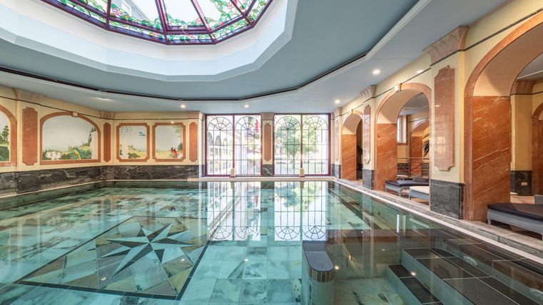Ein luxuriöses Schwimmbad mit einem eleganten Glastdach. Die Wände sind mit kunstvollen Gemälden dekoriert und der Raum ist hell und einladend.