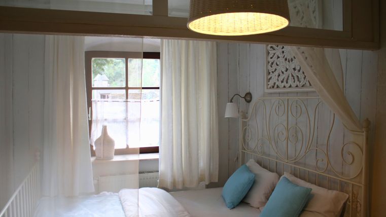 Ein helles Schlafzimmer mit einem weißen Bett und blauen Kissen. Große Fenster lassen viel Licht herein und verleihen dem Raum eine gemütliche Atmosphäre.