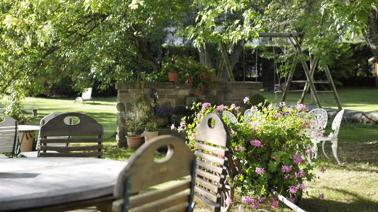 Ein schöner Garten mit Sitzgelegenheiten und blühenden Pflanzen. Im Hintergrund sind Bäume und eine Schaukel zu sehen.