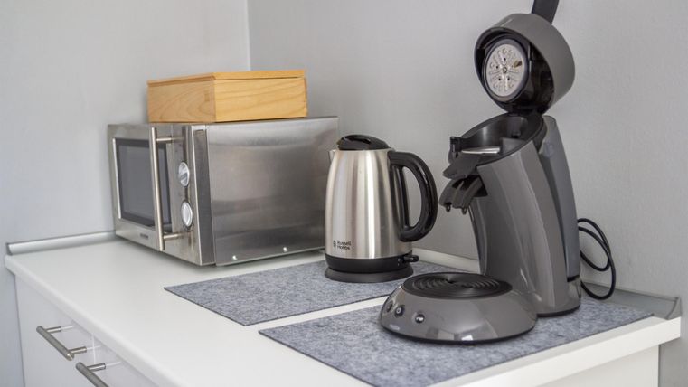 Eine moderne Küchenzeile mit einem Wasserkocher, einer Kaffeemaschine und einer Mikrowelle. Die Küchengeräte sind ordentlich auf einem weißen Tisch angeordnet.