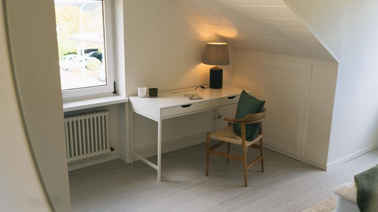 Ein heller Arbeitsplatz mit einem weißen Schreibtisch und einem Stuhl steht unter einem Fenster. Eine Lampe sorgt für gemütliches Licht in dem Raum.