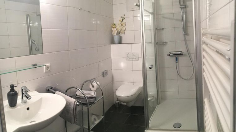 Ein modernes Badezimmer mit einer Dusche, einem Waschbecken und einer Toilette. Die Wände sind weiß gefliest, und es gibt eine Pflanze auf dem Regal.