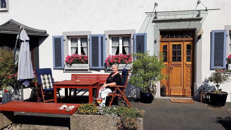 Eine Frau sitzt entspannt auf einer Terrasse vor einem schönen Haus mit blauen Fensterläden und blühenden Pflanzen. Der Tisch und die Stühle aus Holz laden zum Verweilen ein.