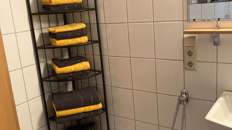 Ein Badezimmer mit weißen Fliesen und einem Handtuchregal. Das Regal ist mit gelben und grauen Handtüchern gefüllt.