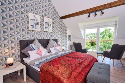 Ein gemütliches Schlafzimmer mit einem großen Bett, das mit bunten Kissen geschmückt ist. Der Raum hat eine moderne Wandgestaltung und große Fenster mit Blick ins Grüne.