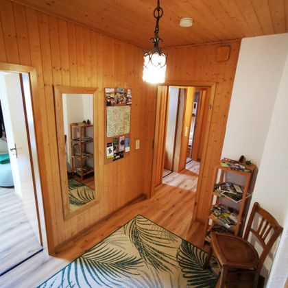 Ein gemütlicher Flur mit Holzverkleidung und einem großen Spiegel. Links steht ein Stuhl und rechts befindet sich ein Regal mit Zeitschriften.