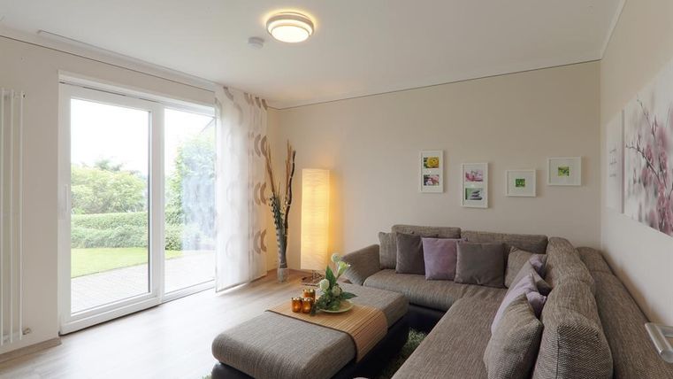 Ein modernes Wohnzimmer mit einem grauen Sofa und einem gemütlichen Teppich. Große Fenster lassen viel Tageslicht herein und bieten einen Blick ins Grüne.