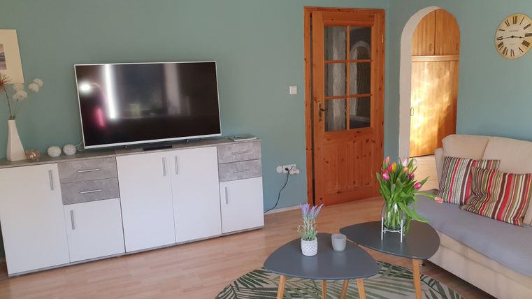 Ein gemütliches Wohnzimmer mit einem Fernseher, einer hellen Couch und einem Tisch. Der Raum hat grüne Wände und Pflanzen auf dem Tisch, die eine einladende Atmosphäre schaffen.