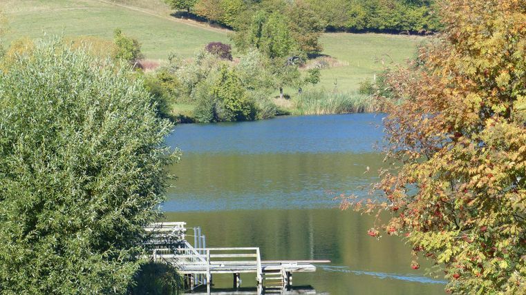Ein ruhiger See umgeben von Bäumen und sanften Hügeln. Eine Holzterrasse lädt zum Entspannen ein.