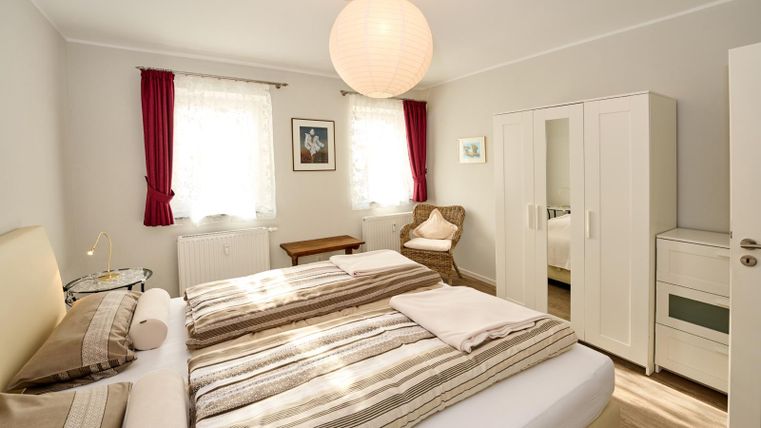 Ein helles Schlafzimmer mit einem großen Bett und gestreiften Bettwäsche. Es gibt Fenster mit weißen Vorhängen und einen gemütlichen Sessel in der Ecke.