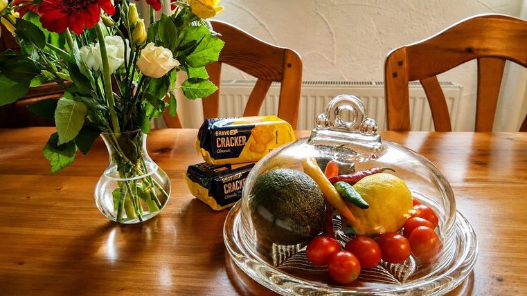 Ein Holztisch mit einer Glasglocke, unter der sich Tomaten, eine Avocado und eine Zitrone befinden. Daneben steht ein Blumengebinde und eine Packung Cracker.