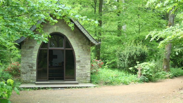 Eine kleine Kapelle im Wald mit einem steinernen Bogen und einem Kreuz daneben.