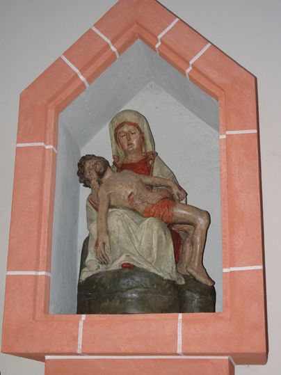 Eine Statue der Pietà zeigt Maria, die den leblosen Körper Jesu hält. Die Skulptur ist in einer Nische platziert und hat einen einfachen, neutralen Hintergrund.