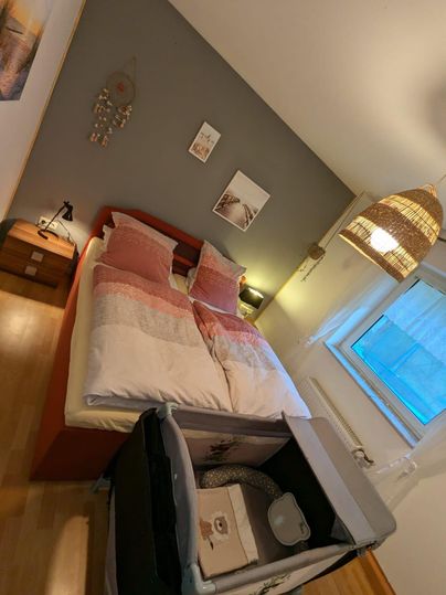Ein modernes Schlafzimmer mit einem Doppelbett und dekorativen Kissen. Im Raum steht auch ein Babybett und ein Fenster mit natürlichem Licht.