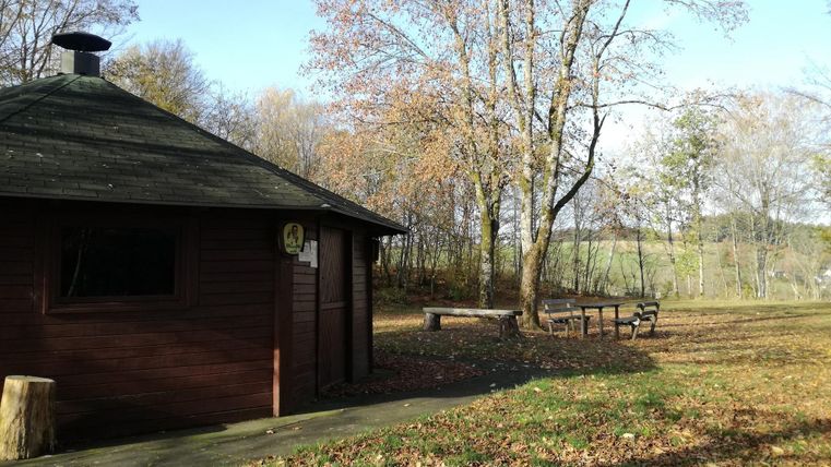Ein kleines Holzhaus umgeben von herbstlichen Bäumen und buntem Laub. Im Vordergrund steht eine Bank auf einem Weg.