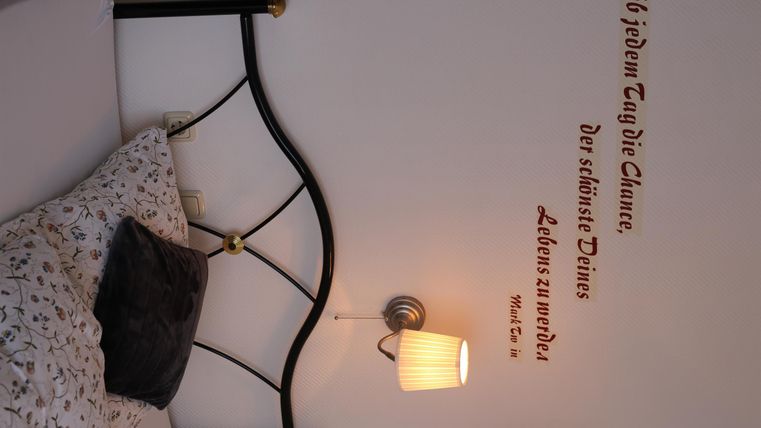 Ein gemütliches Schlafzimmer mit einem Metallbett und einer Wandlampe. An der Wand ist ein inspirierendes Zitat in brauner Schrift zu sehen.