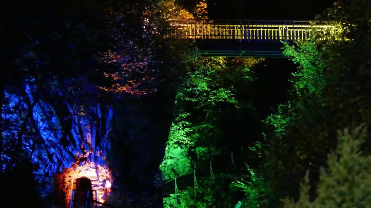 Eine brückenförmige Struktur über einem beleuchteten Felsen in der Nacht. Bunte Lichter sorgen für eine besondere Atmosphäre in der Umgebung.