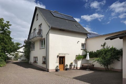 Ein modernes Wohnhaus mit einem Balkon und einem hellen Fassadenanstrich. Der gepflasterte Hof ist von Bäumen und Sträuchern umgeben.