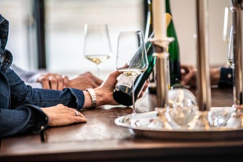 Ein eleganter Tisch mit Gläsern und einer Weinflasche. Hände von Personen sind sichtbar, während sie den Wein einschenken.