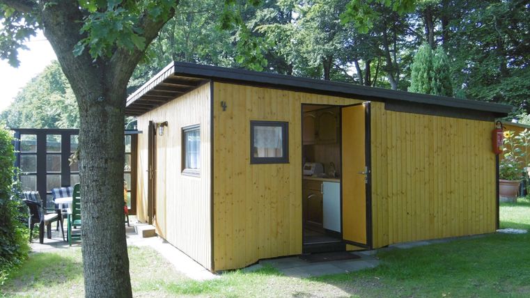 Ein kleines, gelbes Holzhaus steht in einem grünen Garten. Es hat eine offene Tür und ist von Bäumen umgeben.