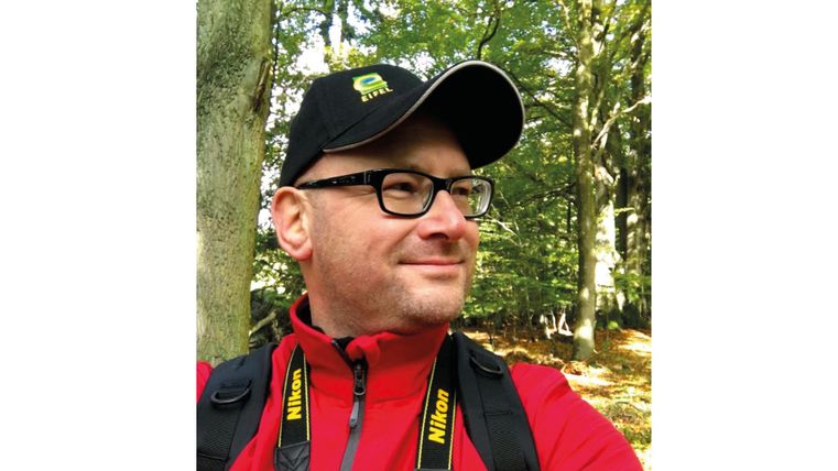 Ein Mann mit Brille trägt ein rotes Jackett und eine schwarze Mütze im Wald. Er schaut in die Kamera und hält eine Kamera um den Hals.