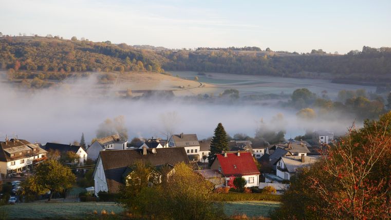 Eine ruhige Landschaft mit einem Dorf im Morgennebel. Sanfte Hügel und grüne Wiesen umgeben die Gebäude.