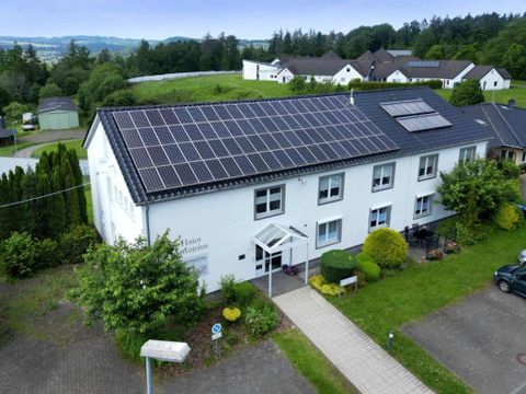 Ein modernes Gebäude mit Solarpanels auf dem Dach. Der Eingangsbereich ist von Pflanzen und Grünflächen umgeben.