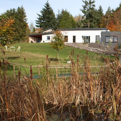 Ein modernes Haus inmitten einer grünen Landschaft. Im Vordergrund befinden sich hohe Pflanzen und eine Sitzgelegenheit.