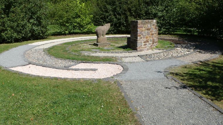 Ein gepflegter Garten mit einem spiralförmigen Weg aus Kies und Steinen. In der Mitte steht ein kleiner Steinblock, umgeben von Pflanzen.