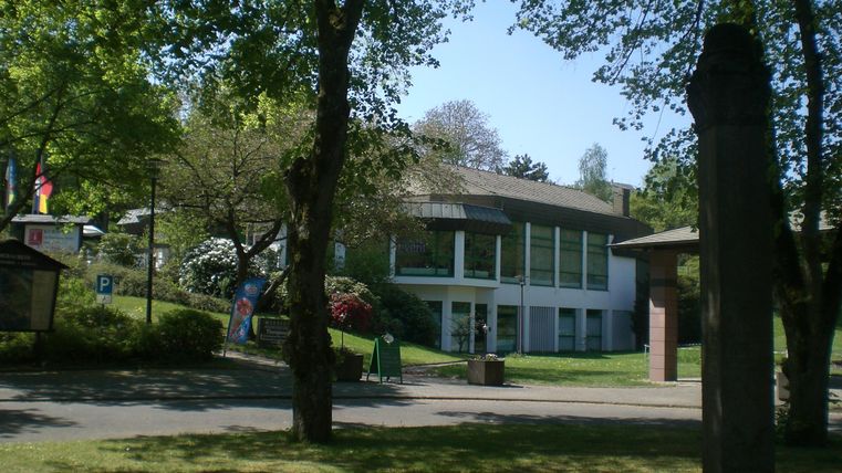A modern building surrounded by trees and green spaces. The sky is blue and it is beautifully sunny.
