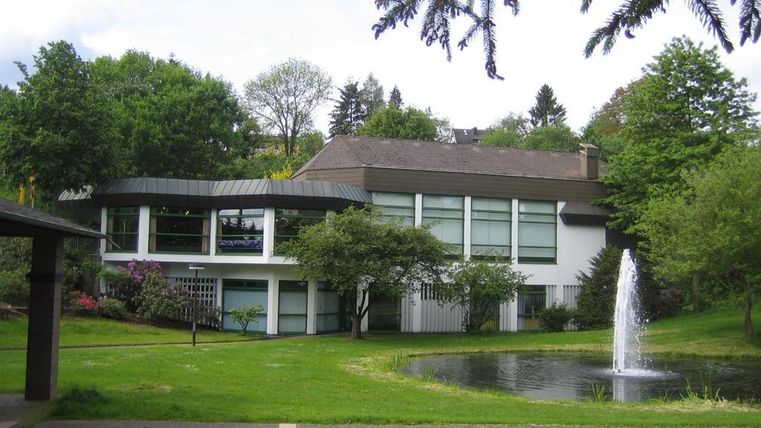 Ein modernes Gebäude umgeben von grünem Landschaftsgarten. Im Vordergrund ist ein kleiner Teich mit einem Wasserfontänen.
