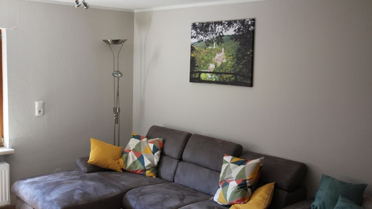 Ein gemütliches Wohnzimmer mit einer großen grauen Couch und bunten Kissen. An der Wand hängt ein Bild von einer grünen Landschaft.
