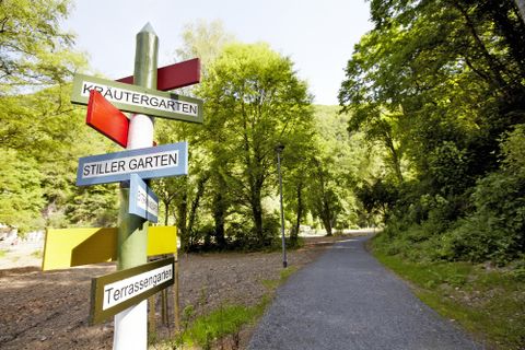 Ein Weg führt durch einen grünen Park, mit verschiedenen Wegweisern, die zu verschiedenen Gartenbereichen führen. Die Umgebung ist ruhig und von Bäumen umgeben.