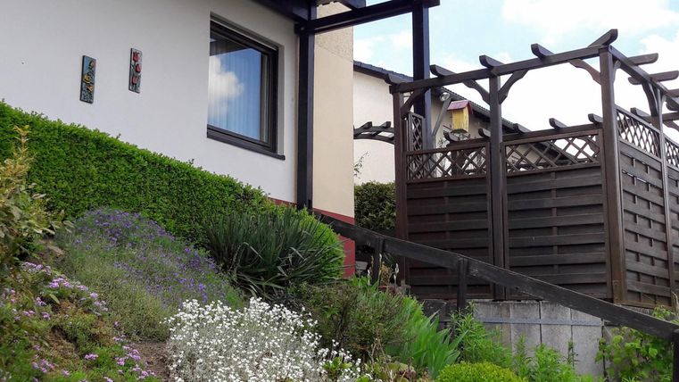Ein modernes Haus mit einem gepflegten Garten und bunten Blumen. Im Hintergrund ist eine Holzterrasse zu sehen.